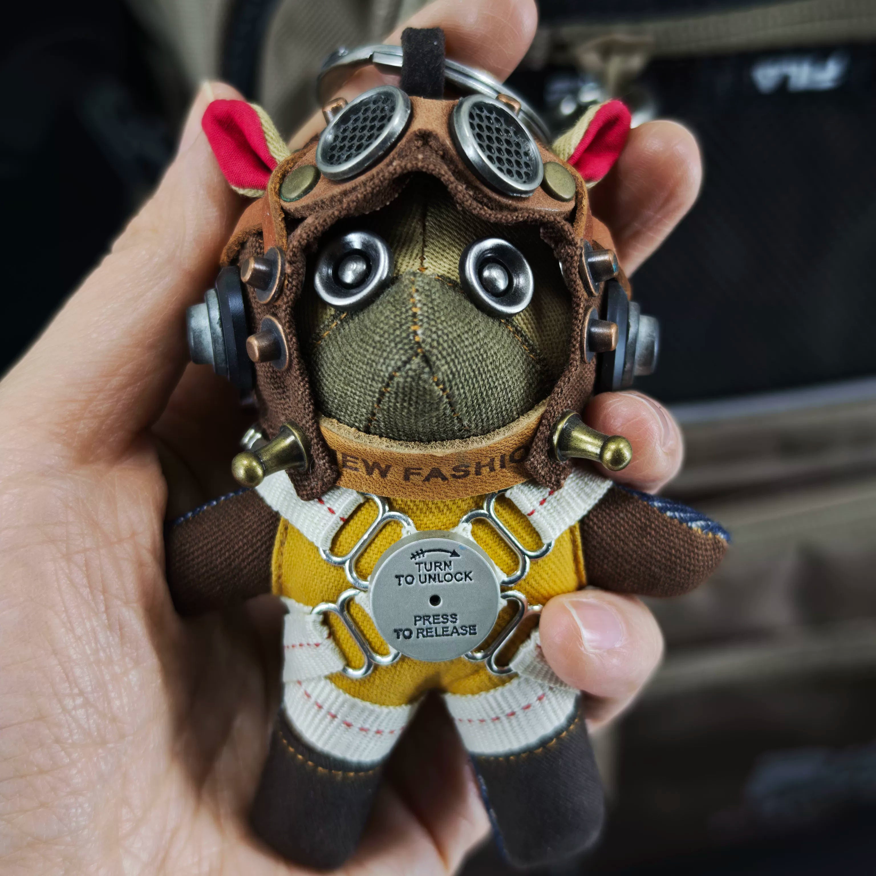 Aviator Pilot Dog Coffee Version — Mini Plush Keychain — backpack clasp view showing waterproof fabric shell, TURN TO UNLOCK metal buckle, PRESS TO RELEASE closure detail — Awhisker