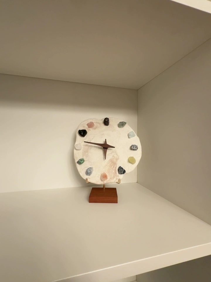 Crystal mineral clock on walnut wood base, close view showing multicolor raw crystal hour markers embedded in white clay disc, brass star hands, shelf display.