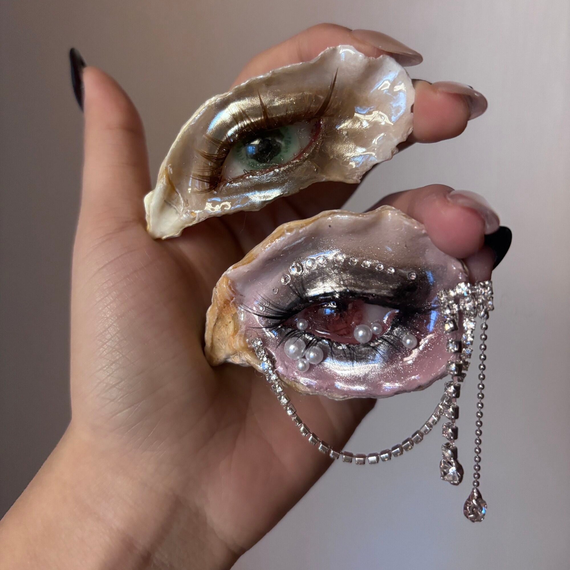 A hand wearing an oyster shell eye brooch with a green iris, brown eyelashes, and pearl accents, shown alongside a second shell with a green iris against a beige background
