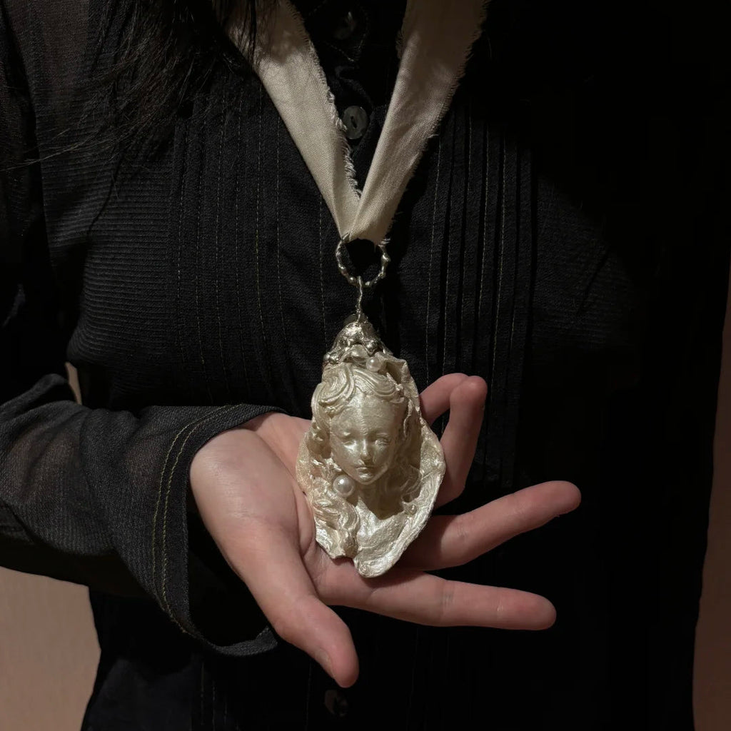 A sculptural silver-toned pendant with a serene woman's face, pearl accents, and an ornate textured metal ring, suspended from a white frayed fabric ribbon against a dark background