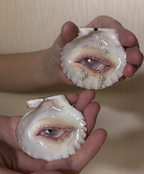 Two hands holding a surreal sculptural piece featuring two scallop shells with realistic eyes and pearl-adorned pearl chains, with a third shell nearby, against a beige striped wallpaper background