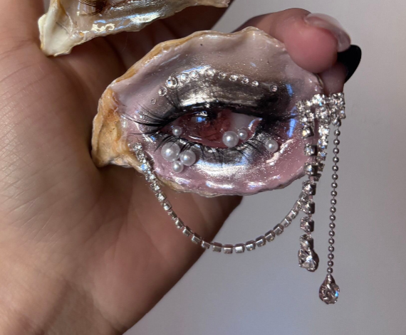 A decorated oyster shell with a red-brown iris, white pearl tears, clear rhinestones along the eyelid, and a silver chain holding a gunmetal crow charm with a red teardrop gem, held by a hand with a burgundy nail