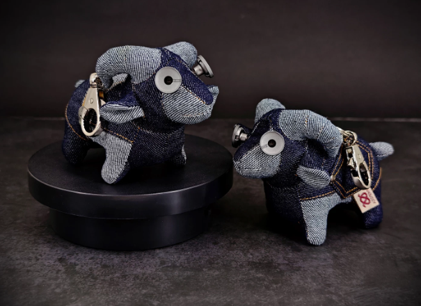 Two denim curled-horn ram plush keychains on a dark surface, patchwork washed-denim bodies with metal snap clasp, yellow collar tag and asymmetric glassy eyes.