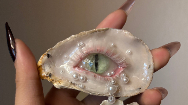 Two oyster shells in pink-purple tones with pearl tears, rhinestones, and dangling gemstone chains displayed on a reflective black surface, showcasing the glamorous iridescent mother-of-pearl texture