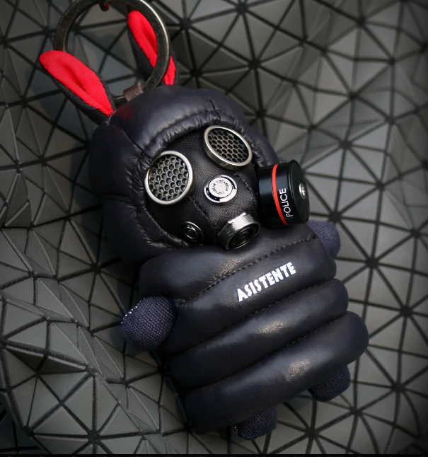 All-black tactical gas mask rabbit plush keychain on a geometric surface, showing triple-filter respirator, red-lined ears, POLICE side canister, ASISTENTE chest plate and metal ring clasp.