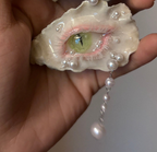 Two oyster shells reimagined as eyes—one with green iris and brown makeup, the other with pink-purple eyeshadow, white pearl tears, rhinestones, and dangling gemstone chains—against a black background with reflective surface