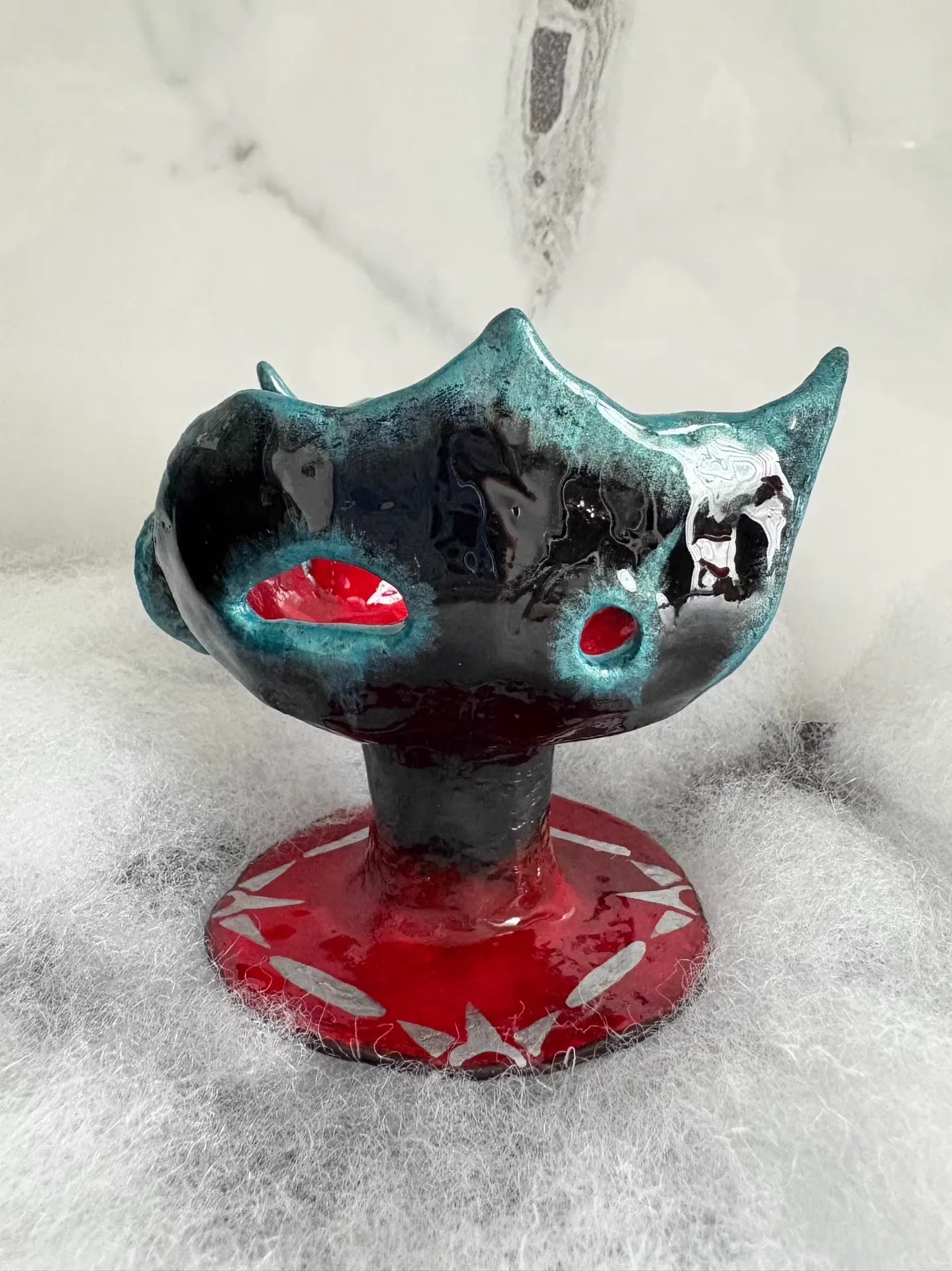 Black and teal glazed alien ceramic vessel, red patterned base, on white fluffy faux fur, marble background