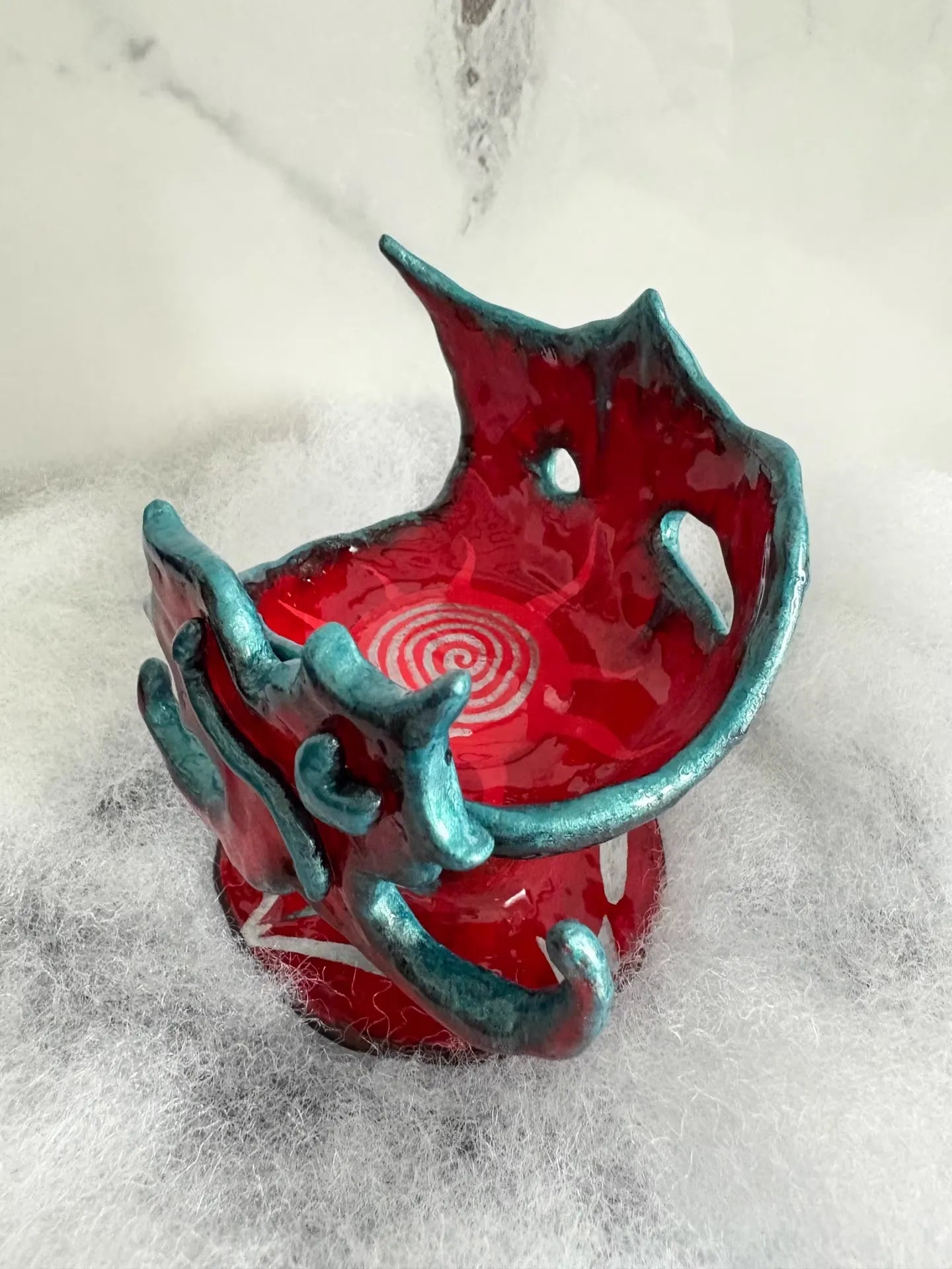 Red glazed alien ceramic vessel with teal trim, white spiral interior pattern, on white fluffy faux fur, marble