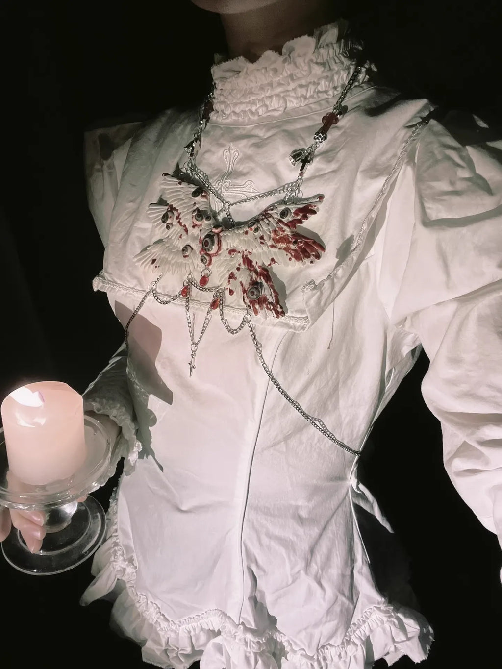 Angel cage eyeball wing body chain on white gothic dress, person holds unlit candle in glass, dark background
