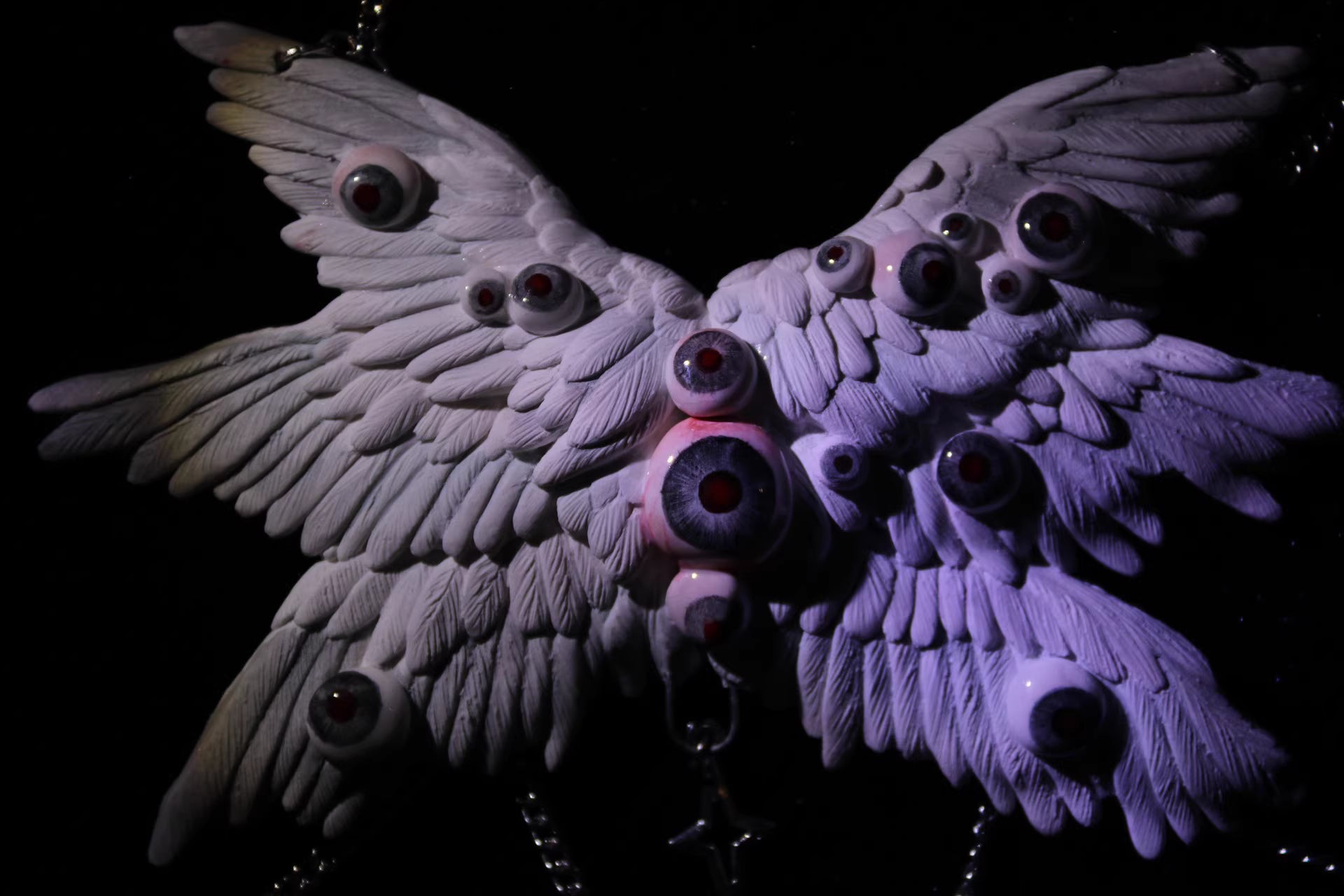 Macabre ceramic wing relic under cold spectral light — the white feather surface shifts toward blue-grey, glass eyeball nodes rendered as pale orbs against the sculpted relief. The chain below is barely visible. The wings remain.