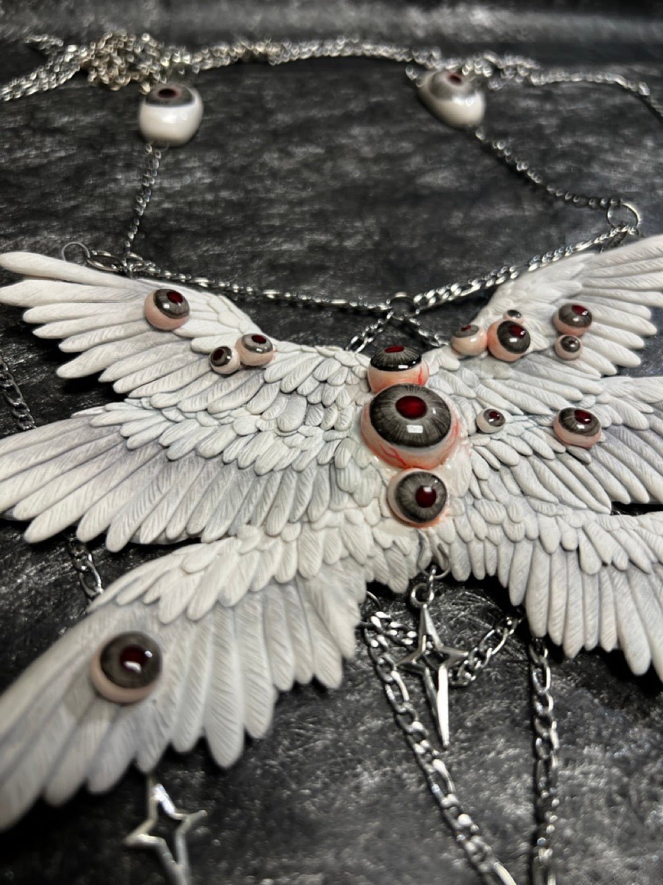 Gothic ceramic body chain artifact displayed front-facing — six white feather wing panels fully extended, glass eyeball inlays distributed across the surface in asymmetric clusters. Silver chain framework visible at lower margin. The spread is total. Nothing is concealed.