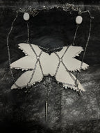 Gothic ceramic angel wing body chain artifact at three-quarter view — white sculpted feather panels with glass eyeball inlays, silver chain structure visible along lower and lateral edges. The feather relief catches directional light, each layer casting a shallow shadow on the one below.