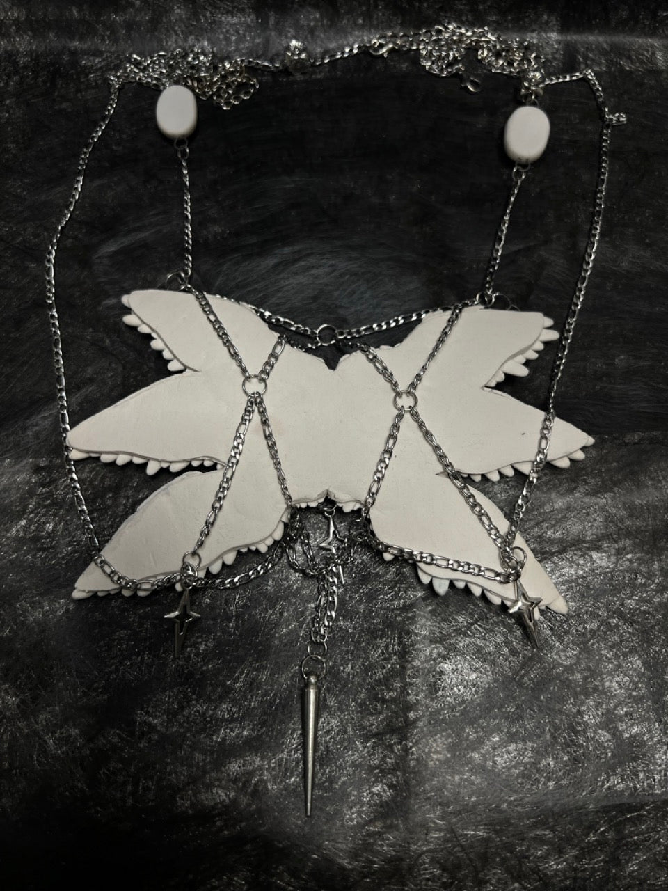 Gothic ceramic angel wing body chain artifact at three-quarter view — white sculpted feather panels with glass eyeball inlays, silver chain structure visible along lower and lateral edges. The feather relief catches directional light, each layer casting a shallow shadow on the one below.