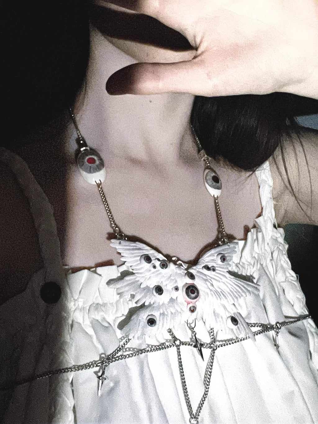 Relic ceramic angel wing body chain in high-contrast monochrome rendering — white feather panels bleached to near-white against deep black ground, glass eyeball inlays visible as dark voids within the wing surface. The chain is a thin silver thread. The form is absolute.