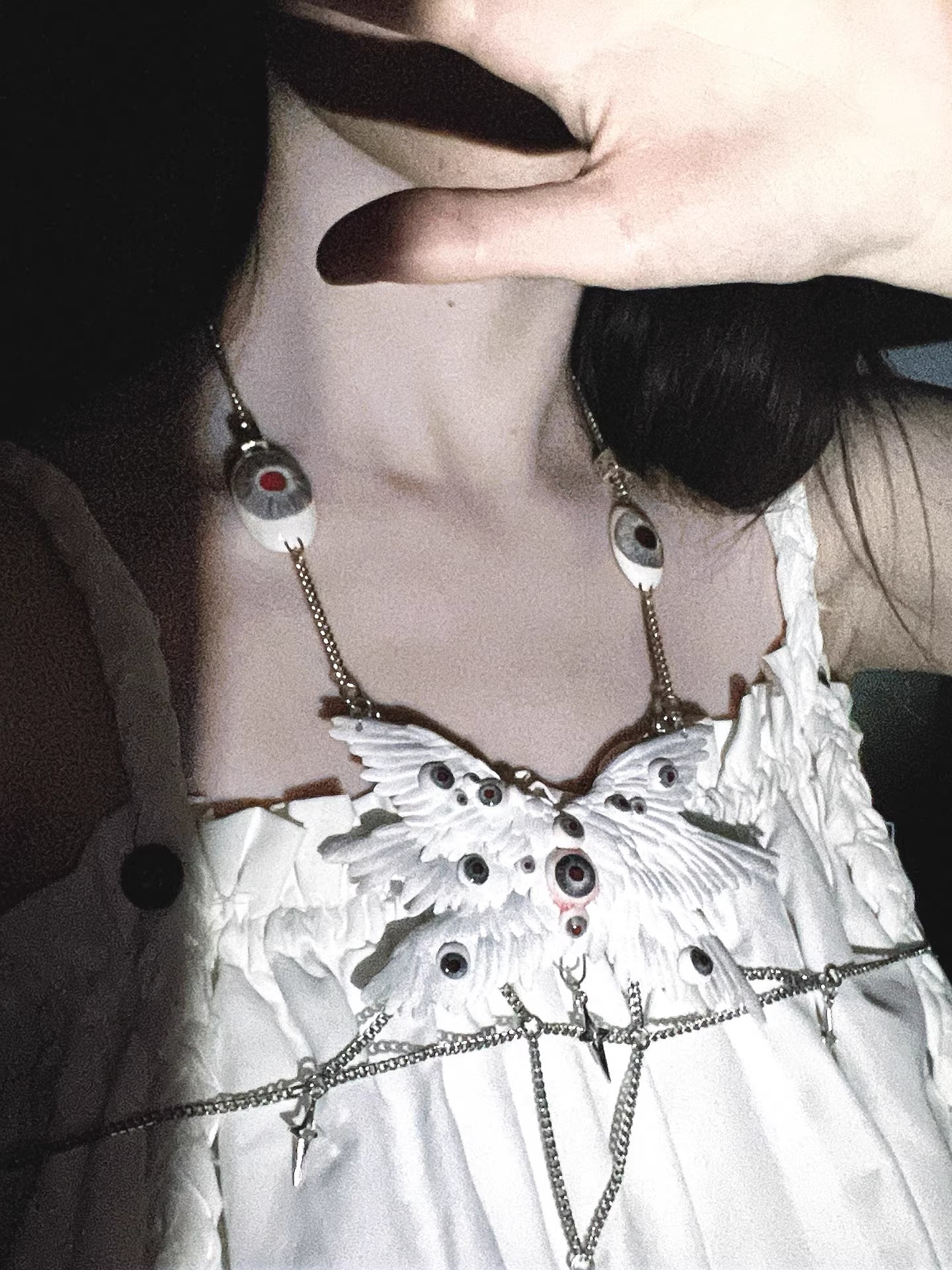 Relic ceramic angel wing body chain in high-contrast monochrome rendering — white feather panels bleached to near-white against deep black ground, glass eyeball inlays visible as dark voids within the wing surface. The chain is a thin silver thread. The form is absolute.