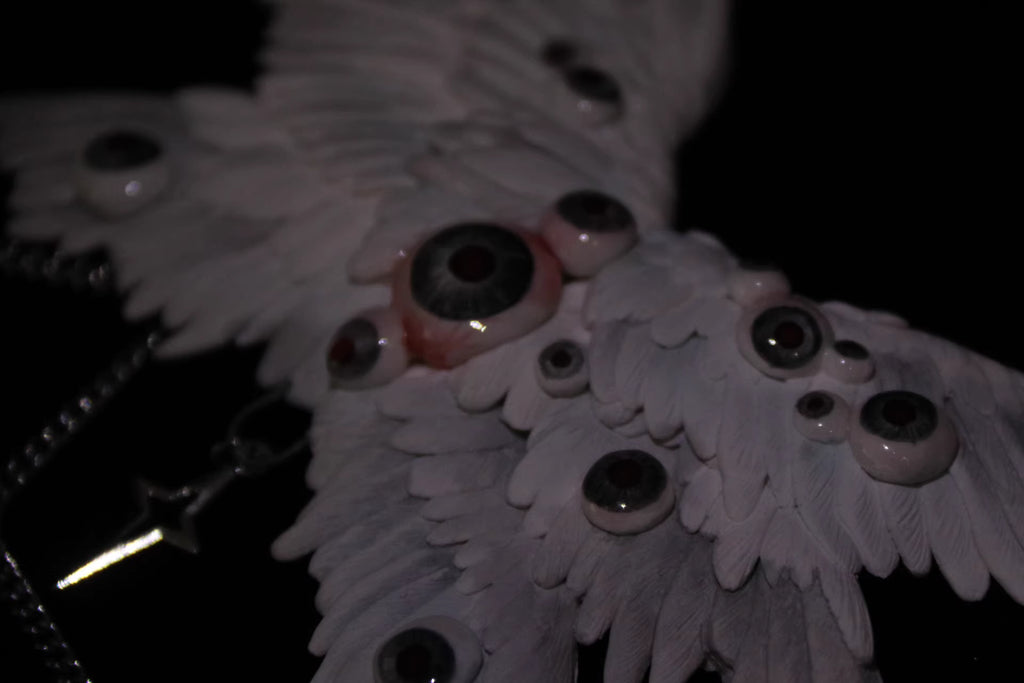 Talisman ceramic angel wing chain relic rendered near-silhouette — rim light traces the outer feather edge against total black, the wing form reduced to its essential geometry. Glass eyeball nodes catch single points of light. The rest is void.