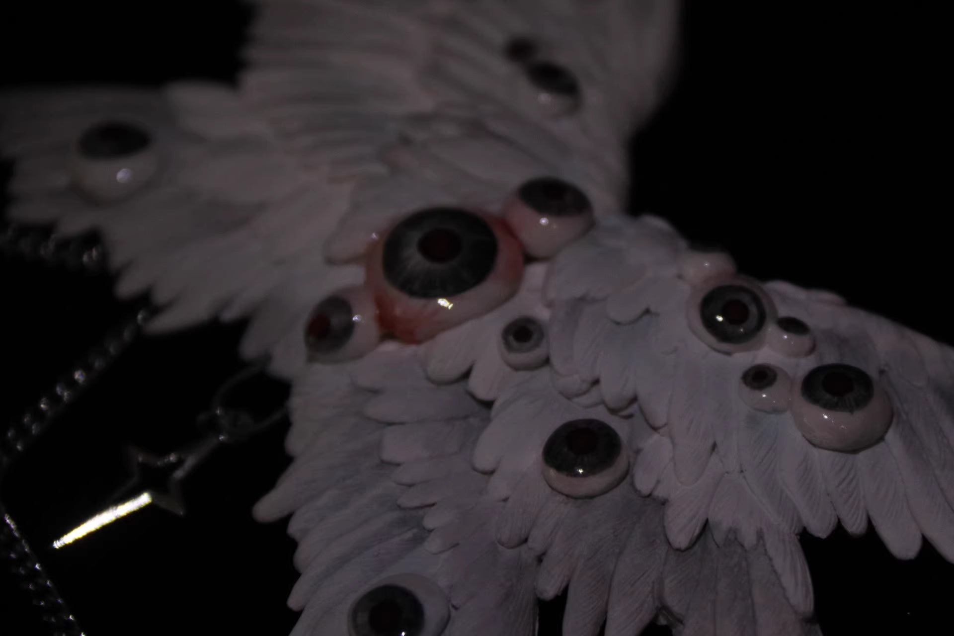 Talisman ceramic angel wing chain relic rendered near-silhouette — rim light traces the outer feather edge against total black, the wing form reduced to its essential geometry. Glass eyeball nodes catch single points of light. The rest is void.