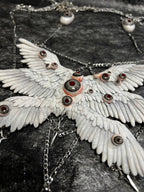 Relic ceramic angel wing body chain captured at oblique angle — white sculpted feather panels with glass eyeball settings, silver chain descending to four-pointed star pendants. Light falls unevenly across the wing surface, deepening the relief between feather layers. The stars hang still.