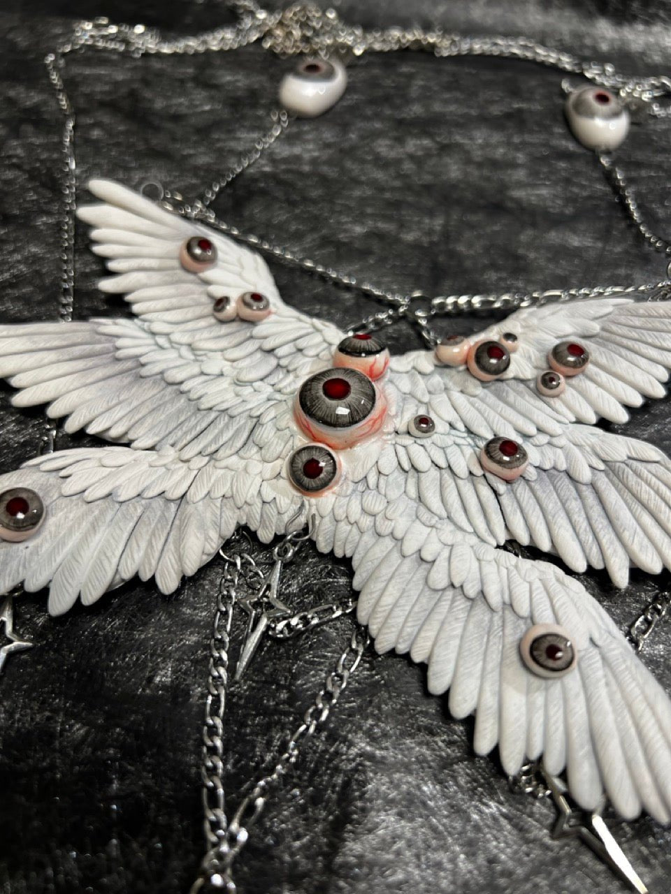 Relic ceramic angel wing body chain captured at oblique angle — white sculpted feather panels with glass eyeball settings, silver chain descending to four-pointed star pendants. Light falls unevenly across the wing surface, deepening the relief between feather layers. The stars hang still.