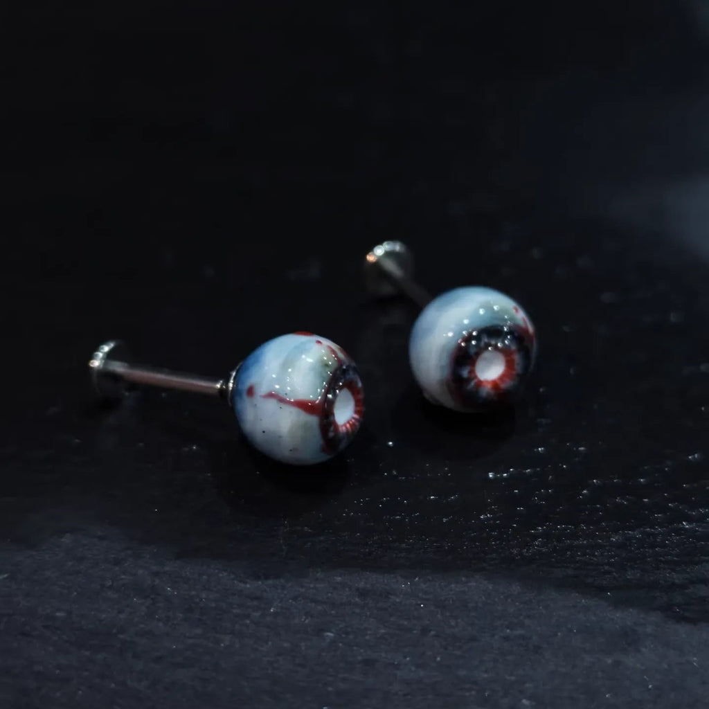 Pair of glossy resin bloodshot necrotic eyeball cartilage studs on dark textured background