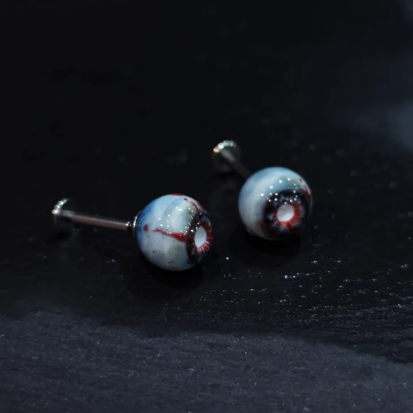 Pair of glossy resin bloodshot necrotic eyeball cartilage studs on dark textured background