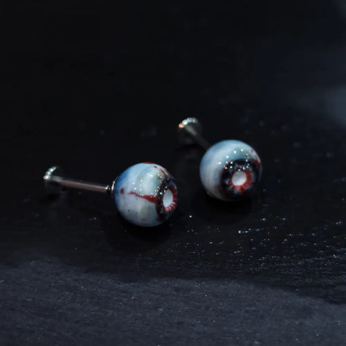 Pair of glossy resin bloodshot necrotic eyeball cartilage studs on dark textured background