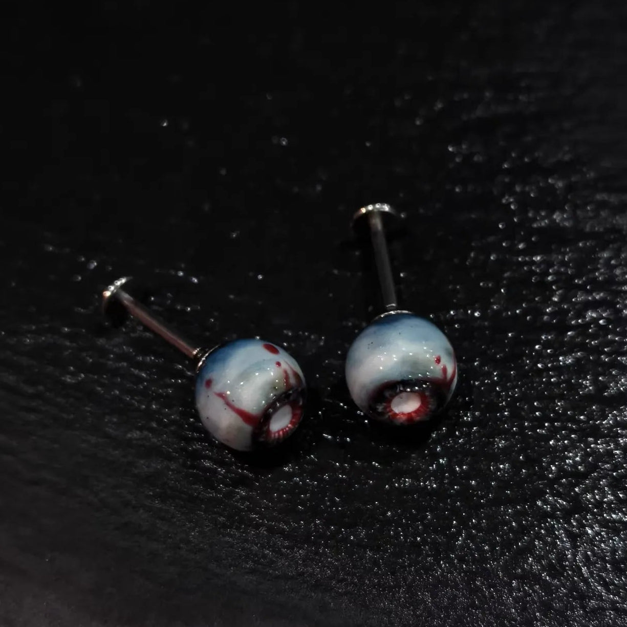 Pair of glossy resin bloodshot necrotic eyeball cartilage studs on solid black background