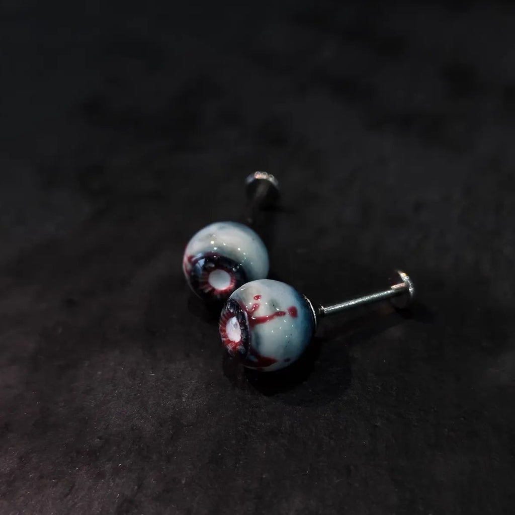 Side view of pair of cinnabar necrotic eyeball cartilage studs on dark sparkly black background