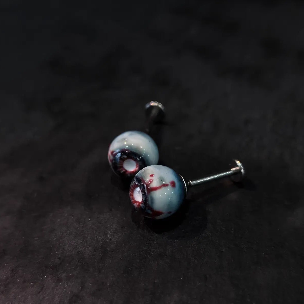 Side view of pair of cinnabar necrotic eyeball cartilage studs on dark sparkly black background