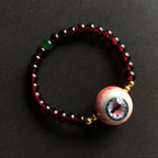 Single strand Cthulhu bracelet, cracked red beads, green accent, gold spacers, focal eyeball on dark background