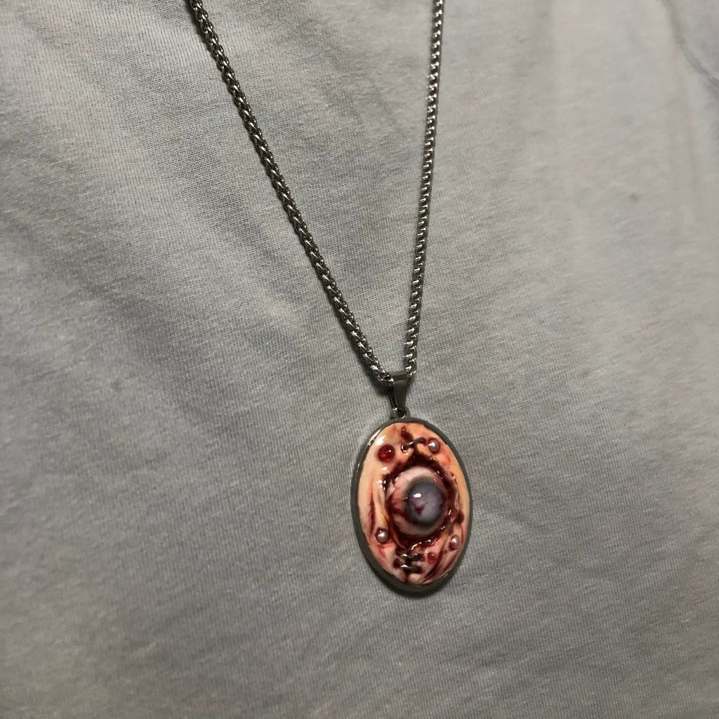 Gothic pendant necklace with raw flesh eyeball design worn on chest, gunmetal chain in V-shape, grey heathered fabric background