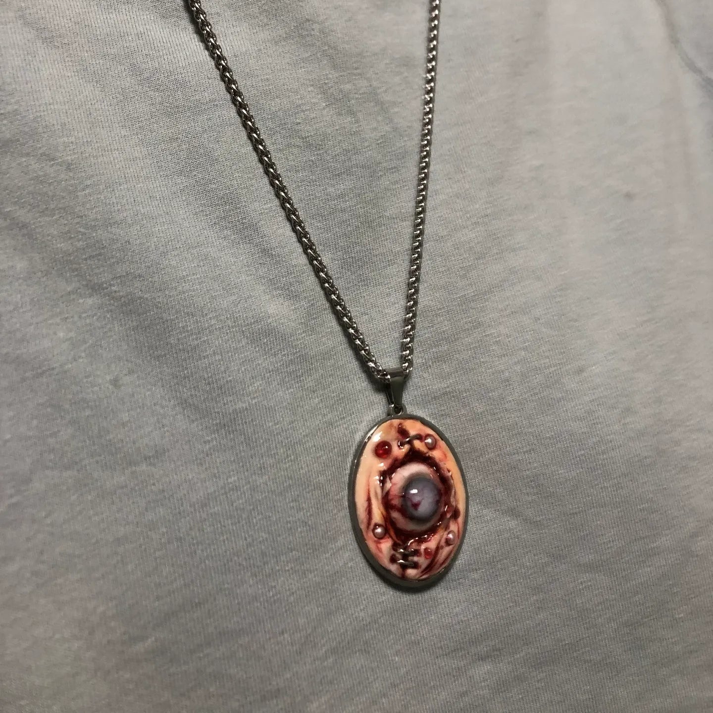 Gothic pendant necklace with raw flesh eyeball design worn on chest, gunmetal chain in V-shape, grey heathered fabric background