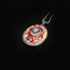 Elevated top-down view of oval pendant with ball chain detail on upper right, marbled flesh surface with red veining on black