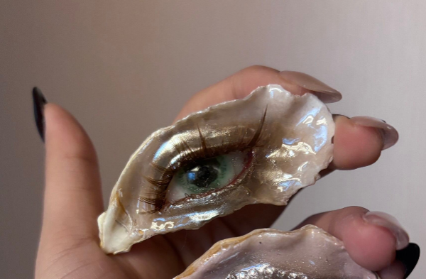 A close-up of an oyster shell with a cat-eye design showing a green iris, vertical slit pupil, pink eyelids, white pearl tears, and a dangling pearl chain on black enamel nail against a grey background