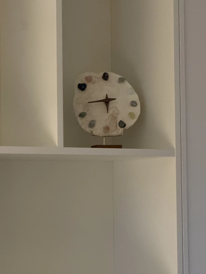 Crystal mineral clock on walnut wood base, hand-formed clay disc with twelve raw gemstone hour markers, brass four-point hands, displayed on white shelf.