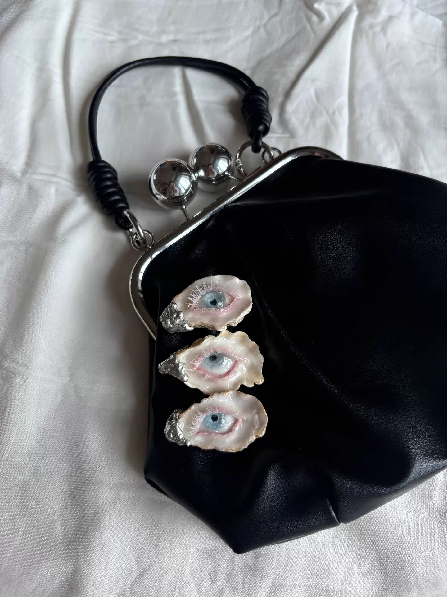 A pebbled black leather kiss-lock purse with a scallop shell eye, realistic eye sculpture, pearl tears, and a silver chain strap, held by a hand against a beige textured wall