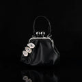 A black leather kiss-lock frame purse with three surreal oyster shell eye embellishments featuring blue irises, pink eyelids, and white eyelashes, silver spherical charms, and a twisted black leather handle against a white fabric background