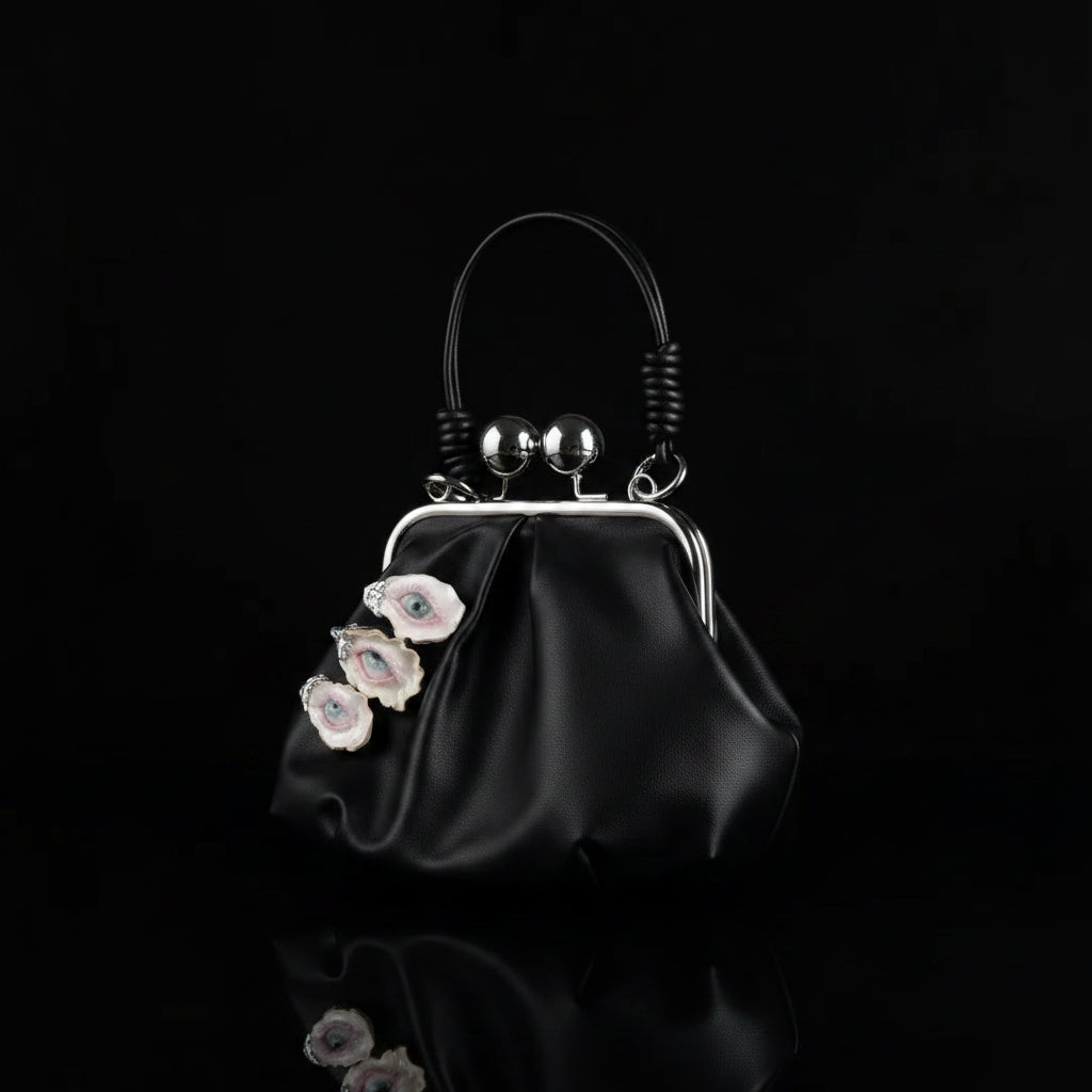 A black leather kiss-lock frame purse with three surreal oyster shell eye embellishments featuring blue irises, pink eyelids, and white eyelashes, silver spherical charms, and a twisted black leather handle against a white fabric background