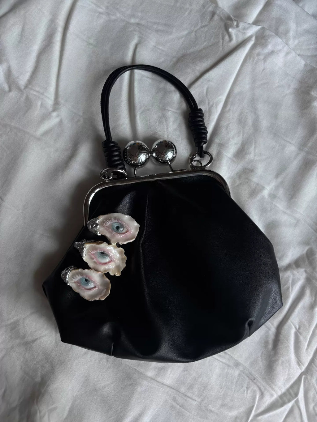 A black leather kiss-lock bag with three blue-eyed shell embellishments and silver clasp balls, shown against a solid black background with a mirror-like reflective surface beneath
