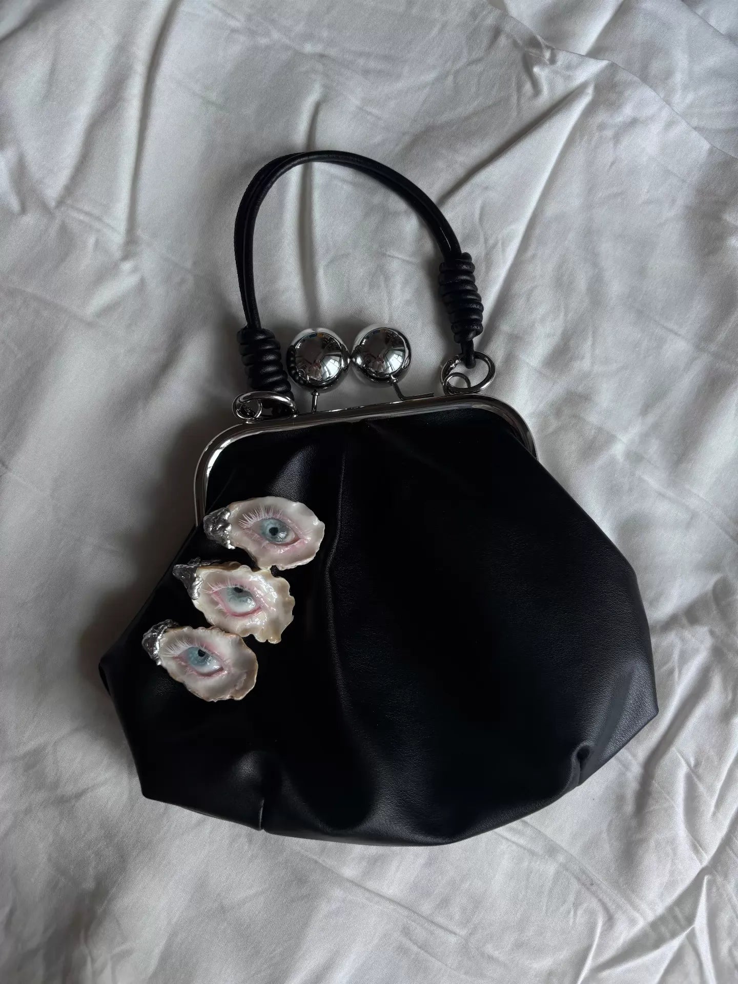 A black leather kiss-lock bag with three blue-eyed shell embellishments and silver clasp balls, shown against a solid black background with a mirror-like reflective surface beneath