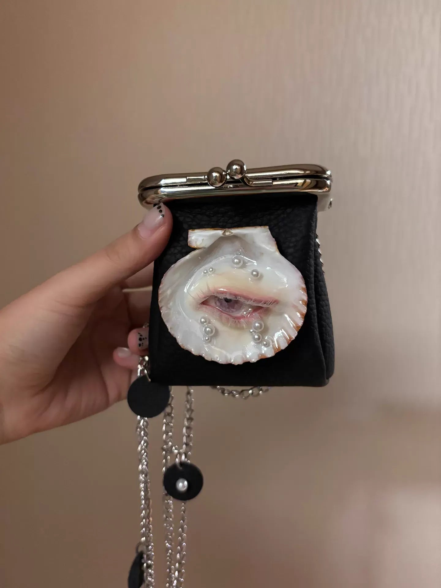 A black leather kiss-lock purse with a scallop shell eye and realistic eye with pearl tears, worn by a person in a black robe and brown shirt against a dark moody background