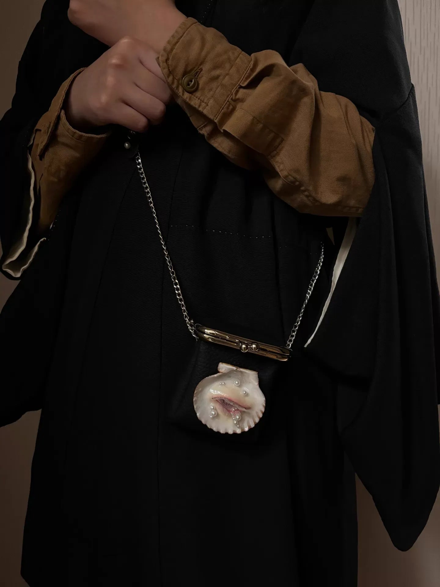 A close-up of a surreal shell eye sculpture with pearl tears and teardrop gemstones on a pebbled leather bag, paired with a scallop shell pendant necklace featuring a sculpted eye on a silver chain