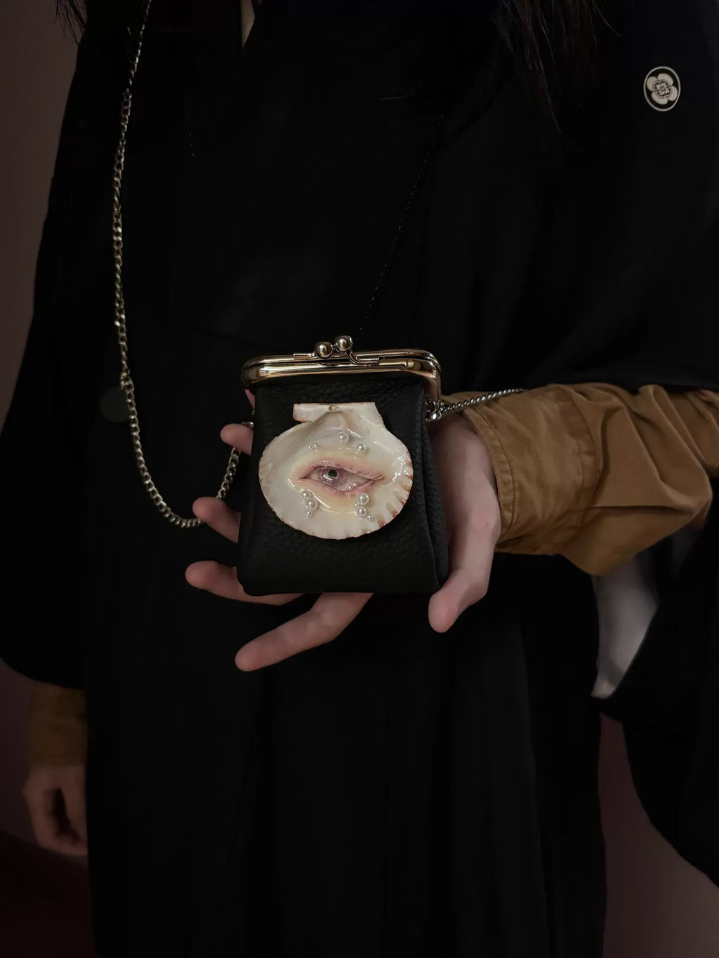 A surreal necklace featuring a sculptural lips-and-pearl design on a scallop shell, paired with a black pebbled leather kiss-lock purse with a shell eye, worn against a dark background