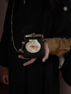 A surreal necklace featuring a sculptural lips-and-pearl design on a scallop shell, paired with a black pebbled leather kiss-lock purse with a shell eye, worn against a dark background