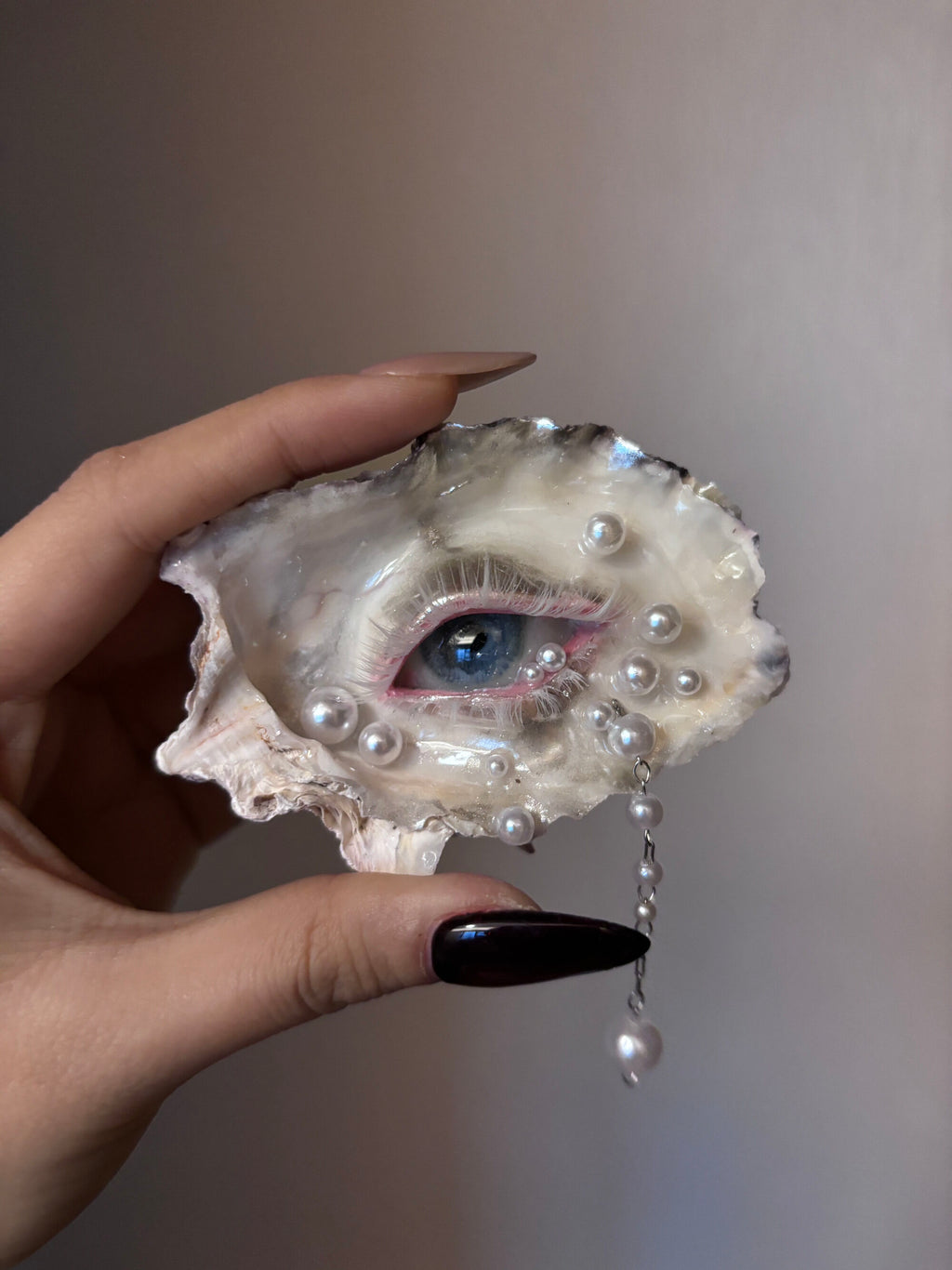 A hand holding an oyster shell transformed into a green-eyed art piece with pink eyelashes, pearl accents, and a dangling pearl chain against a light grey background