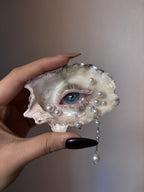 A hand holding an oyster shell transformed into a green-eyed art piece with pink eyelashes, pearl accents, and a dangling pearl chain against a light grey background