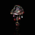 A gothic enamel brooch with a blue-green iris eye, pink eyelashes, snake-like vine border, and dangling iridescent beads, teardrop crystals, pearl, mosaic, and bee charms against a black background