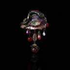 A gothic enamel brooch with a blue-green iris eye, pink eyelashes, snake-like vine border, and dangling iridescent beads, teardrop crystals, pearl, mosaic, and bee charms against a black background