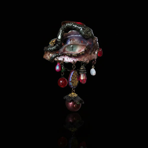 A gothic enamel brooch with a blue-green iris eye, pink eyelashes, snake-like vine border, and dangling iridescent beads, teardrop crystals, pearl, mosaic, and bee charms against a black background