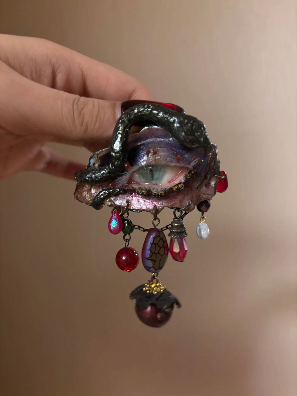 A textured iridescent hair brooch or barrette with a crackled purple-blue bead, red teardrop, glass beads, and a maroon bead with gold cap on a gold-toned chain, held by a hand with dark nail polish